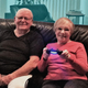 Karen Klein and her husband Bruce smile as they sit together on their sofa with a Playstation 4 controller in hand.
